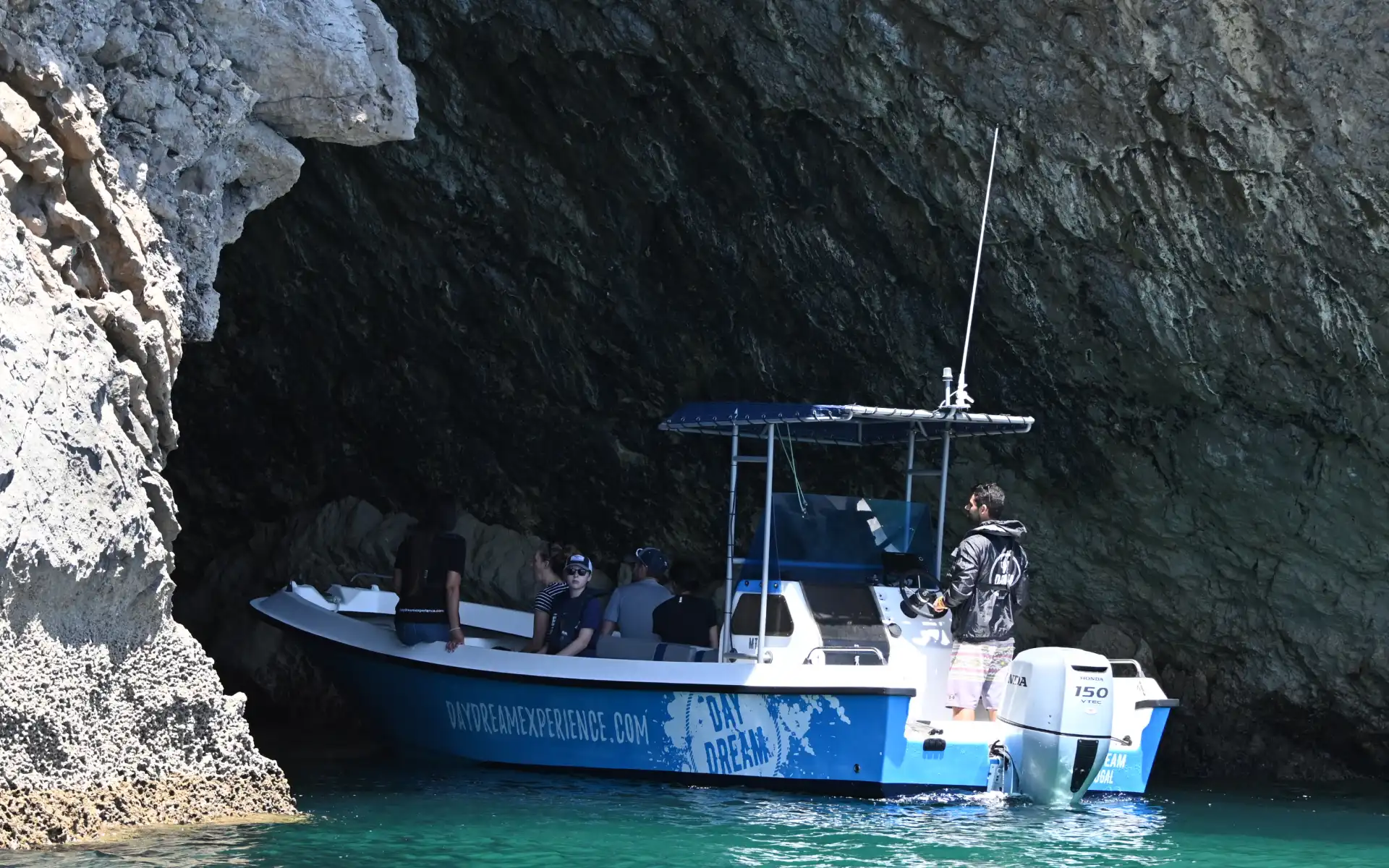 Tourists in a small motorboat exploring a dark sea cave along a rugged limestone cliff on a sunny day.