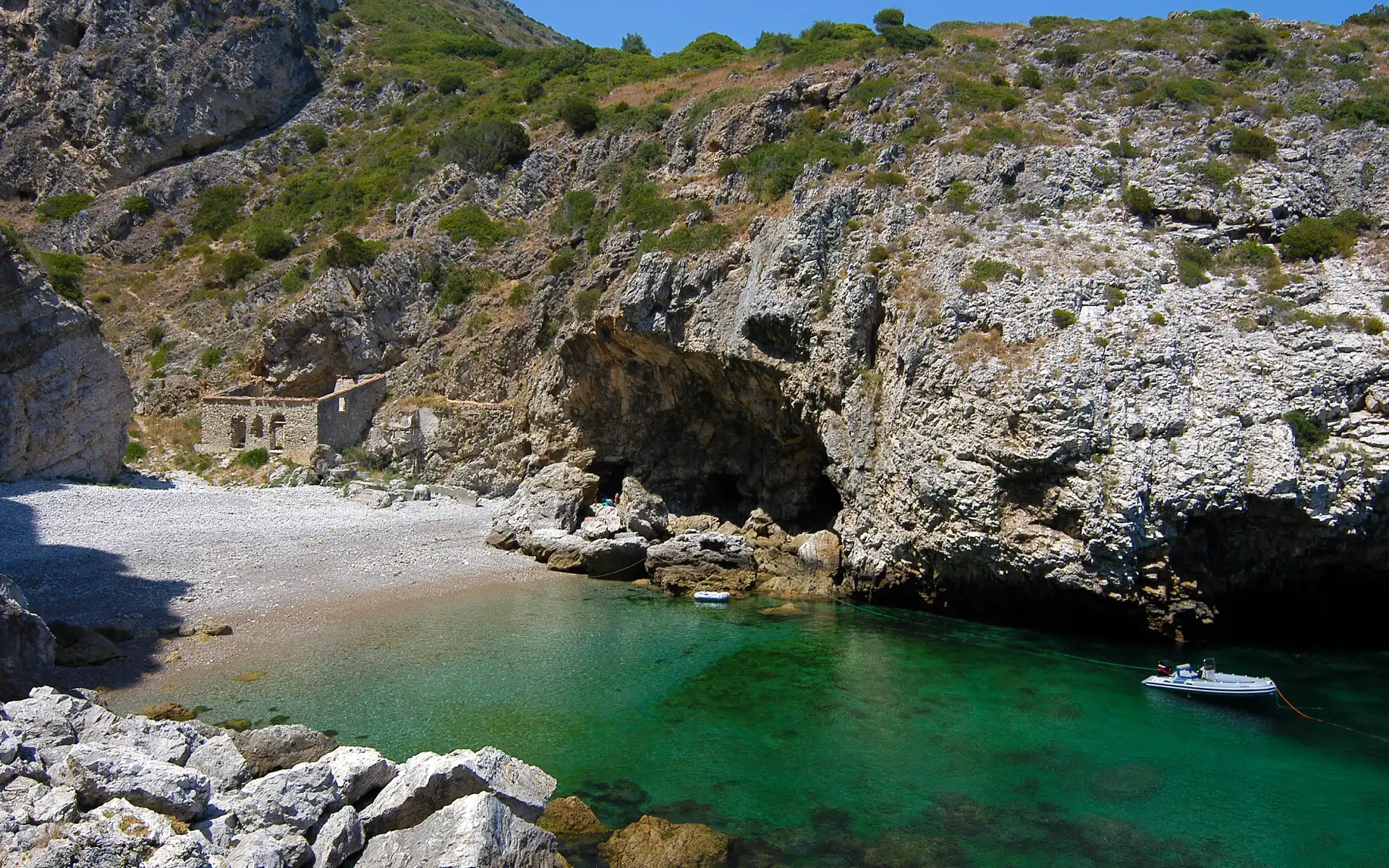 Remote pebbled beach with turquoise waters and a small abandoned stone building nestled among steep rocky cliffs near Sesimbra, Portugal.