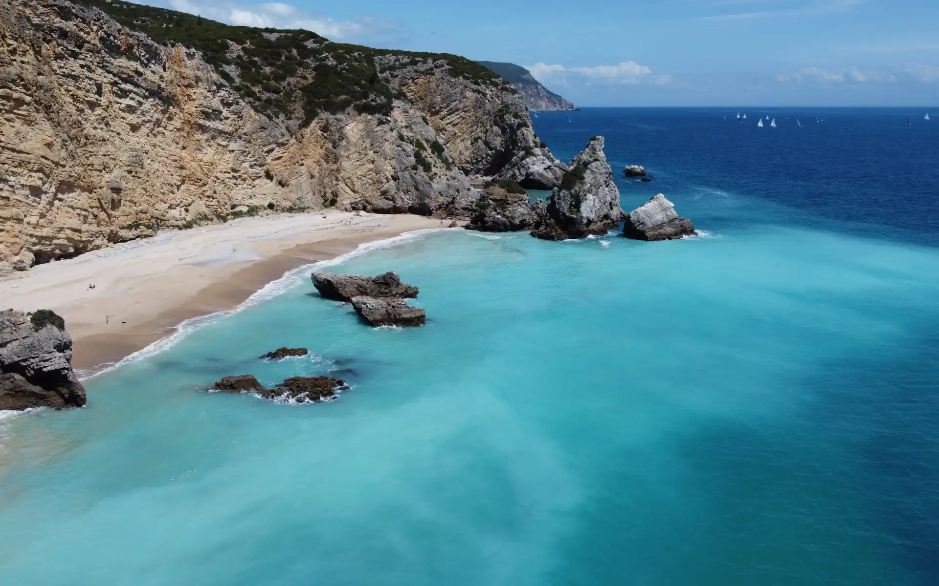 A secluded beach with turquoise waters and rugged rock formations along the shore, backed by steep cliffs covered in greenery under a clear blue sky.