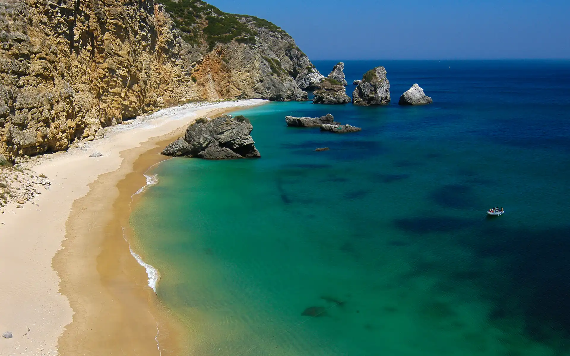 A golden sandy beach with crystal-clear emerald waters, bordered by dramatic rocky cliffs and large rock formations jutting out of the sea under a bright blue sky.