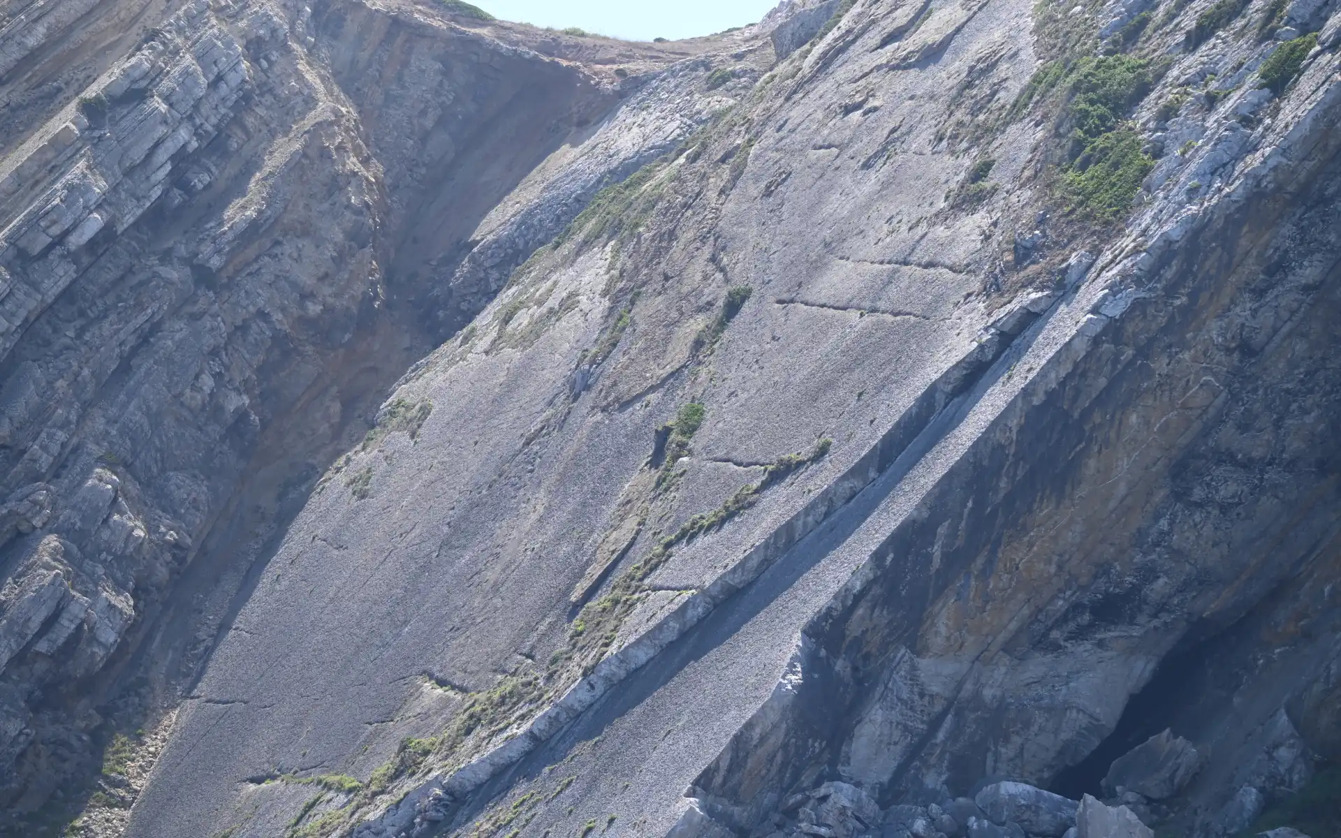 A dramatic limestone canyon or gorge with steep stratified rock walls showing distinct geological layers, scattered green vegetation growing on the cliff faces, and rocky debris at the bottom under an overcast sky.