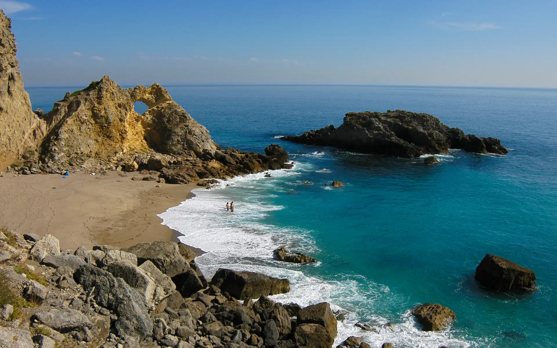 Rocky shoreline with turquoise waves washing up on a secluded sandy beach, where two people wade in the surf beneath clear blue skies.