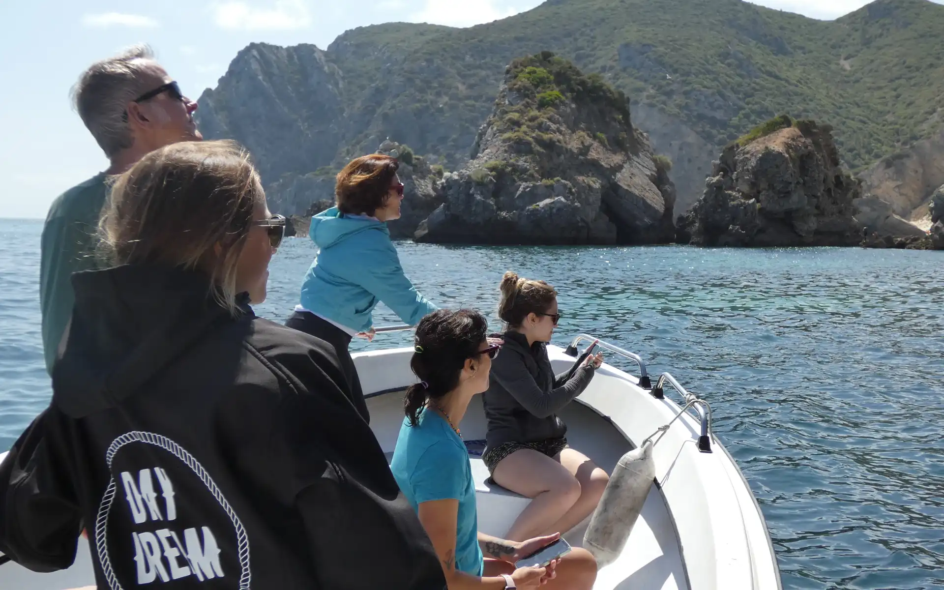 Group of people and a guide on a boat tour observing the coastline and surrounding nature in calm waters, with rugged cliffs and green hills in the background.