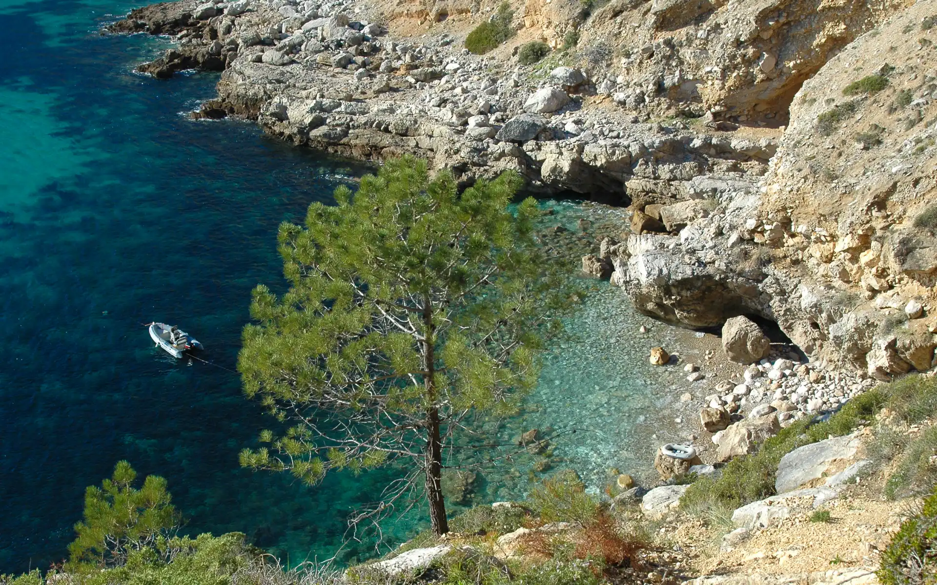 Small inflatable boat anchored near a rocky shore, seen from above through pine trees and clear blue-green waters.
