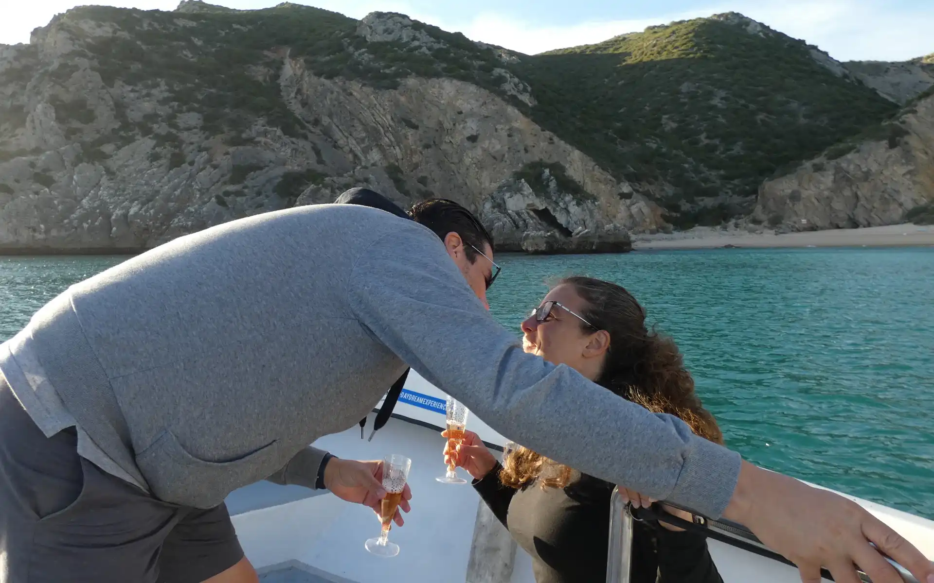 Romantic couple clinking glasses of sparkling wine on a boat, with cliffs and sea in the background.