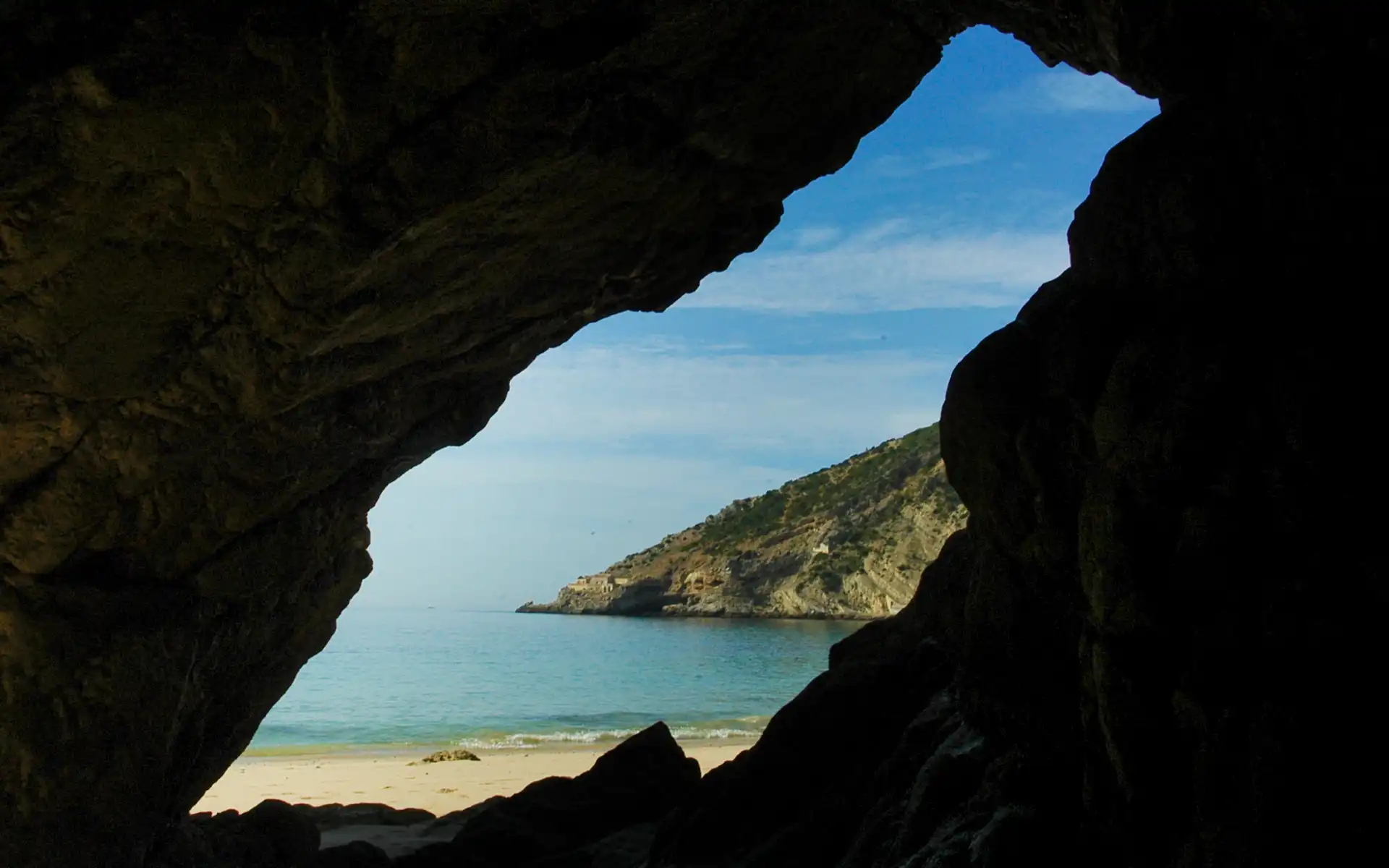View from inside a sea cave looking out to a quiet beach and rocky cliffs under a blue sky.