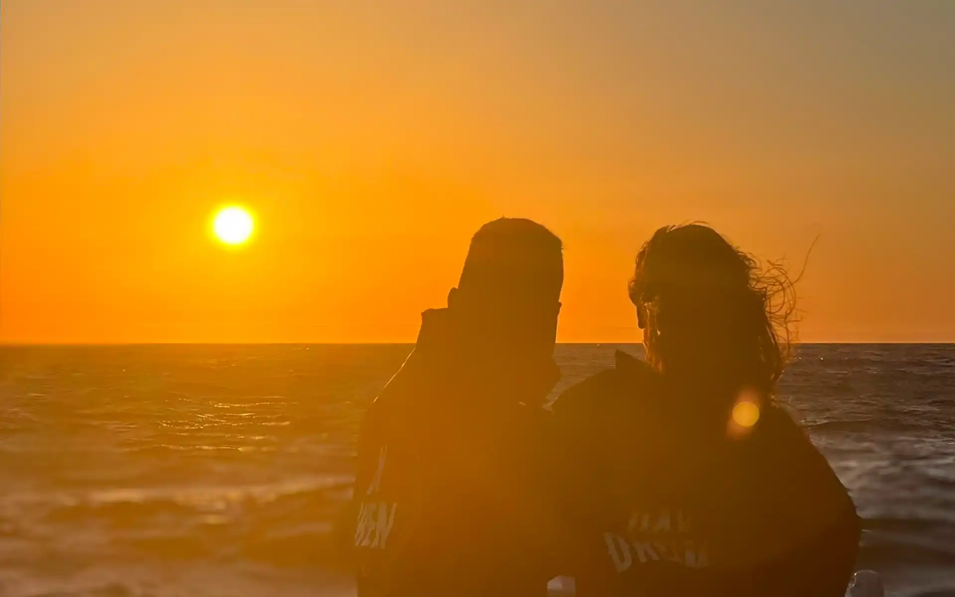 Silhouetted couple on a boat at sunset, bathed in golden light with the ocean and glowing horizon in the background.