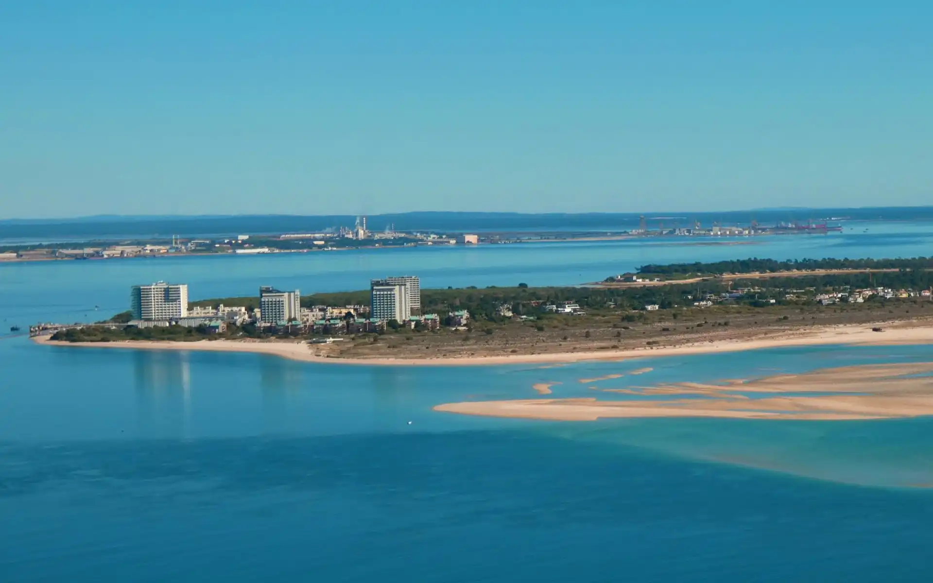 Aerial view of Tróia Island with sandy beaches, modern beachfront buildings, and calm blue waters on a clear day.
